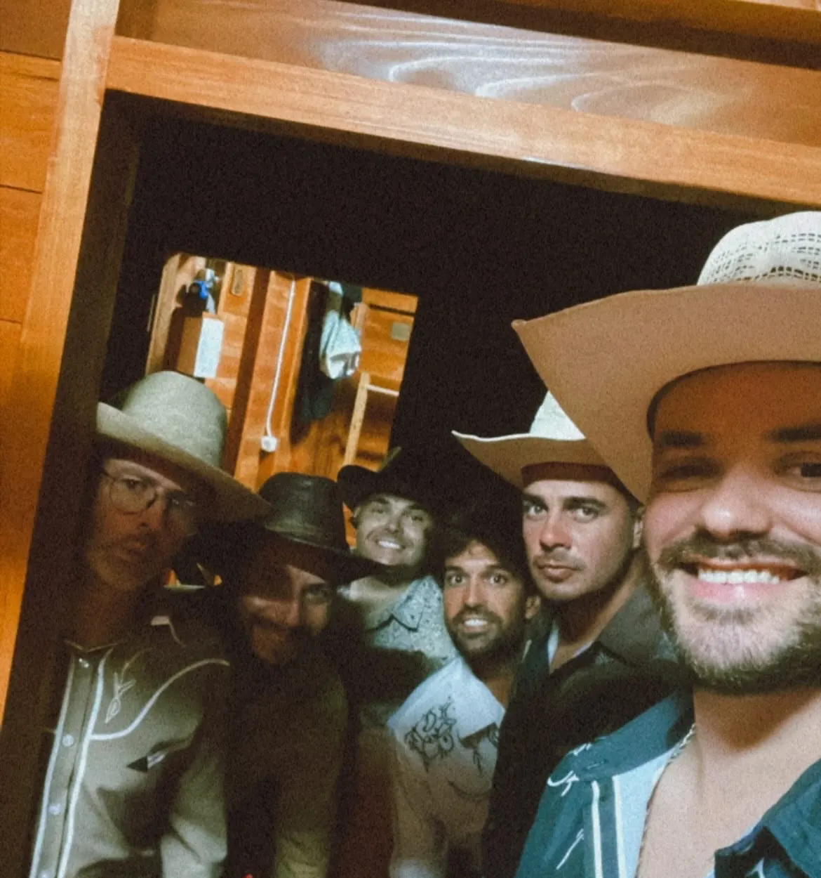 Selfie nos bastidores: seis elementos da banda, de chapéu de cowboy e camisas western, à porta de uma cabana de madeira.