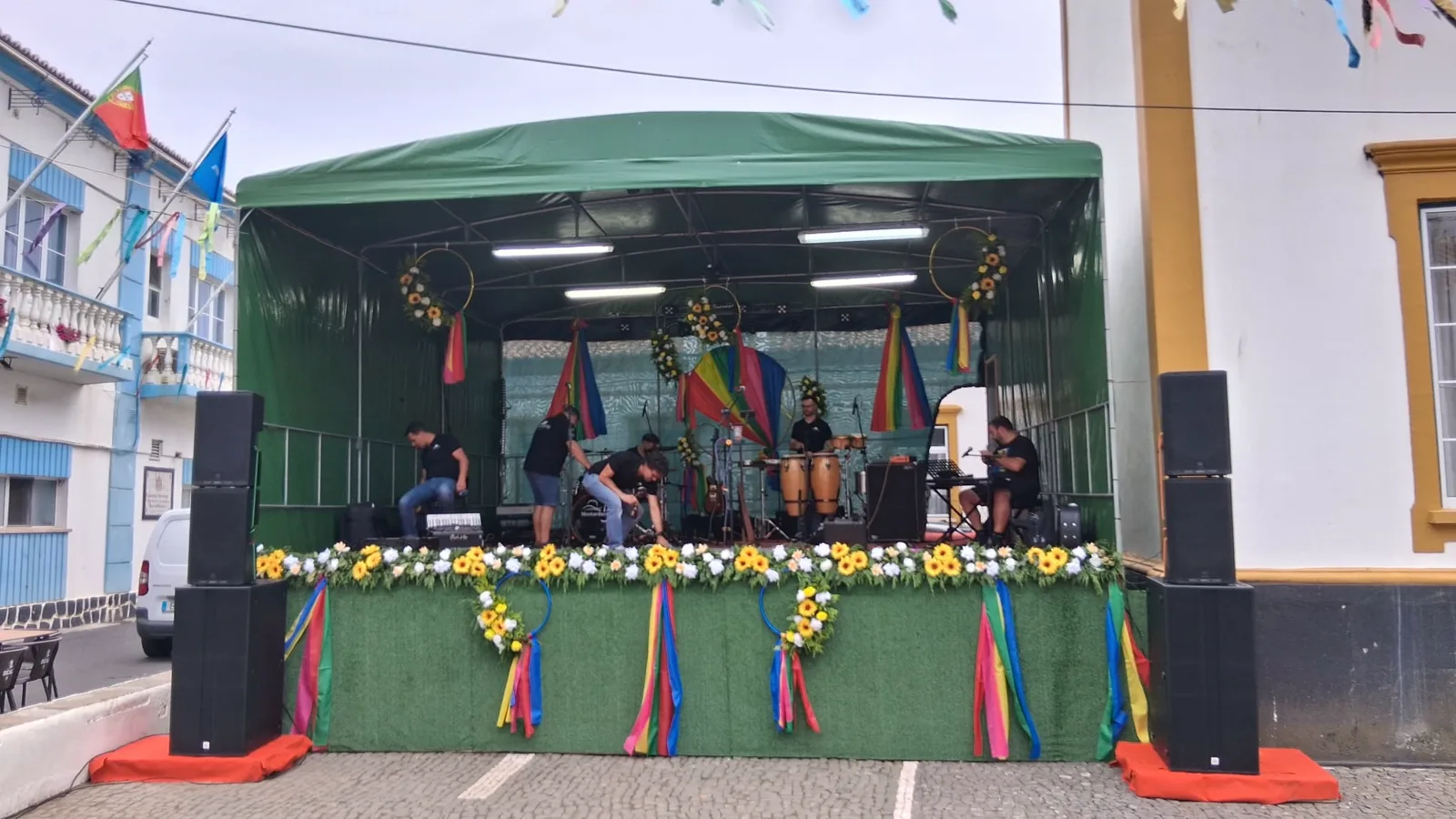 Palco ao ar livre decorado com girassóis e fitas coloridas, com a banda a preparar os instrumentos.